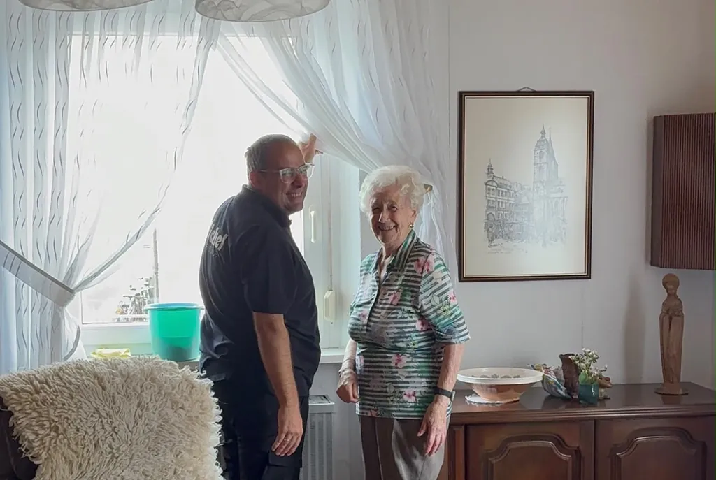 Ein Mitarbeiter von Michel Dienstleistungen hängt mit einer lächelnden Seniorin in ihrem Wohnzimmer die Gardinen auf